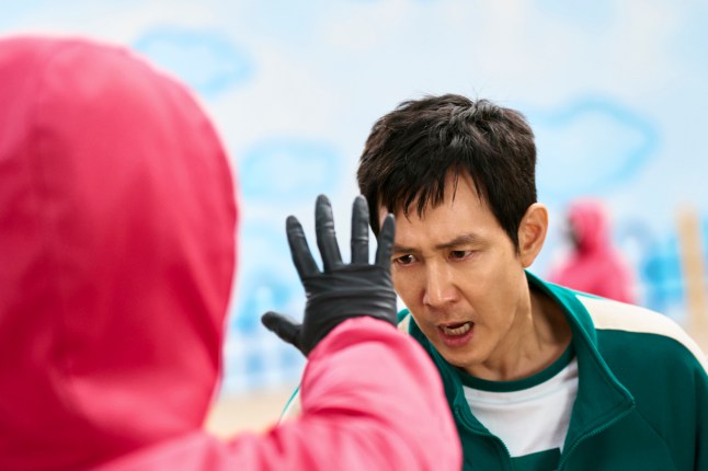 Lee Byung-hun as Hwang In-ho in Squid Game season two, looking at the outstretched hand of a member of staff from who is wearing a pink hooded jumpsuit.