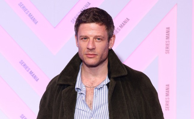 James Norton attends a screening wearing a brown jacket and a blue striped shirt against a pink stamp and repeat.