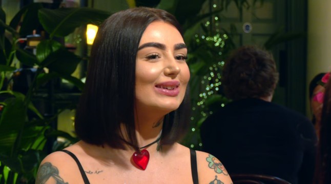 A close up of Emily on First Dates. She is wearing a black strappy top and a necklace with a large red heart pendant.