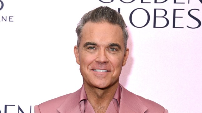 Robbie Williams smiles while wearing a pink suit on a red carpet.