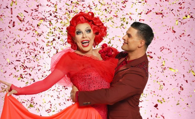 La Voix and Aljaž Skorjanec in a promo shot for Strictly Come Dancing. Pink background. La Voix in red dress and Aljaž in red suit.
