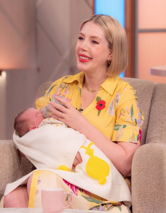 Katherine Ryan holding her new born baby, on This Morning. Wearing yellow dress and cradling baby wrapped in white blanket with yellow design.