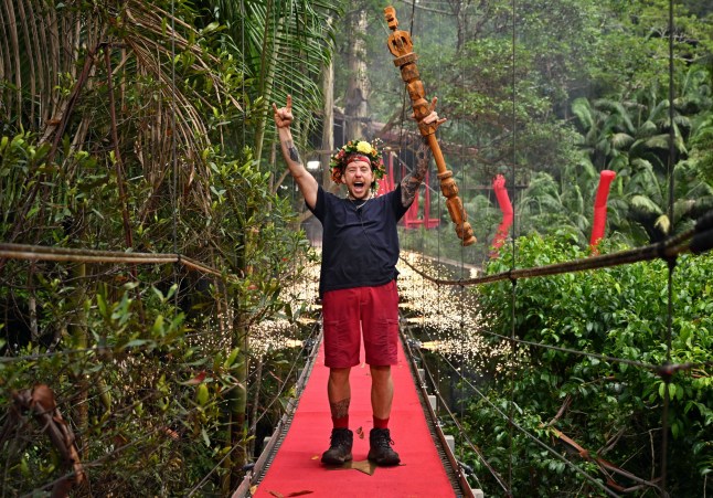 Danny Jones raising his arms in celebration of winning I'm A Celebrity 2024, holding a wooden staff and wearing a jungle crown