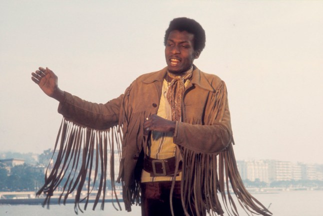 Jimmy Cliff dancing in a suede brown jacket with tassels, in front of a body of water.